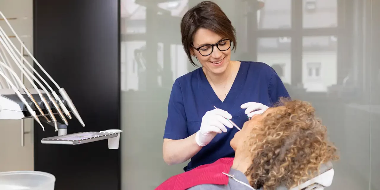 Drau Dr. Mairock überprüft lächelnd das Gebiss einer Patientin auf dem Zahnarztstuhl.